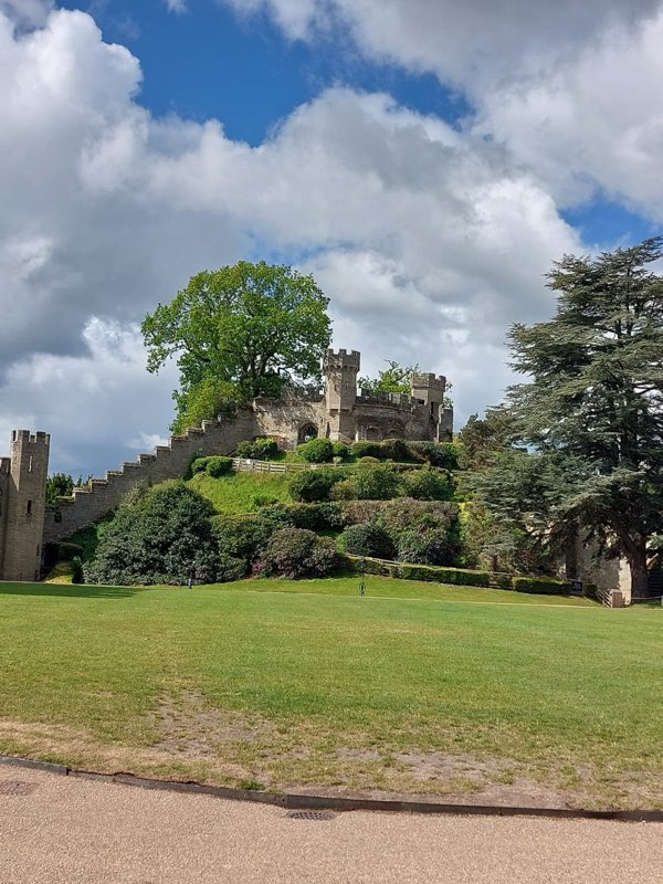 Wide mown lawn, shrub beds and mature trees below castle walls under a cloudy sky