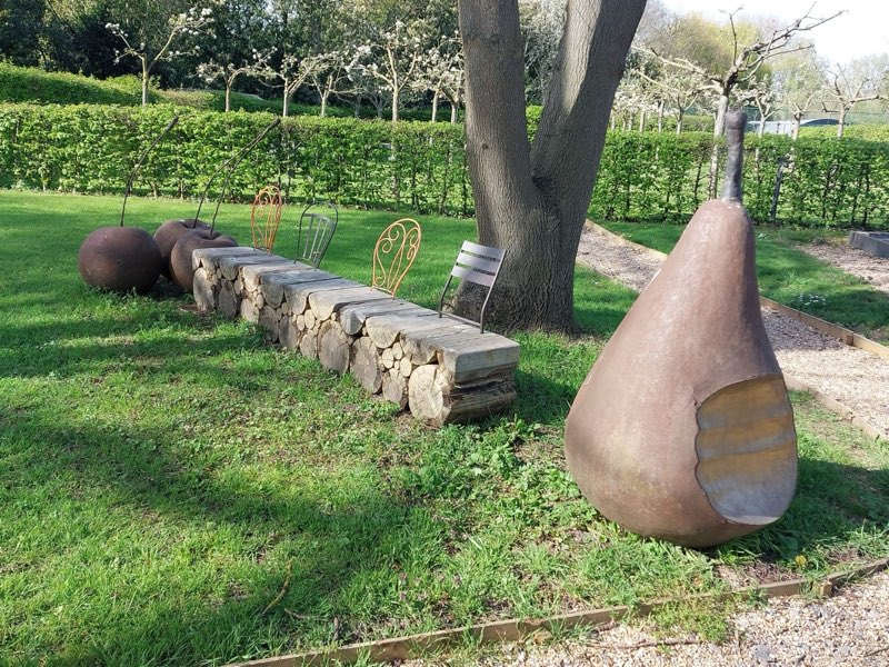 Formal garden with clipped hedges, a log bench and sculptures of fruit, with a blossoming orchard behind